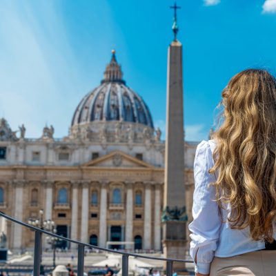 View of the blonde woman from behind in Vatican City. Saint Peter Square with multiple tourists, Saint Peter Basilica and the Vatican Obelisk on the background